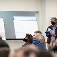 Long shot of audience and mentor in background wearing masks reading abstract book.